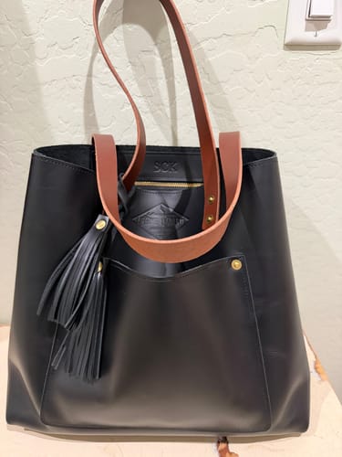 Reviewer's black leather Lifetime Tote with brown handles and a matching black tassel, standing on a surface.