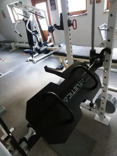 Rezensent zeigt das Ersatzteil, ein schwarzes Trittblech für die Beinpresse, montiert an einer weißen Multipresse in einem Fitnessraum.