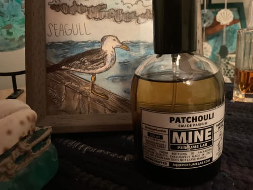 Flacone dell'Eau de Parfum PATCHOULI di un cliente, su una superficie scura accanto a un quadro.