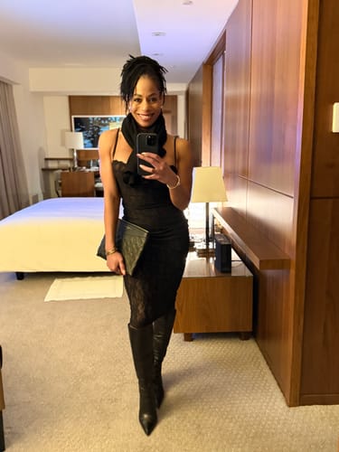 Reviewer taking a mirror selfie in a hotel room, wearing the black Stretch Lace Midi Dress with a scarf and boots.
