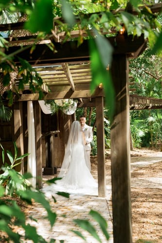 Reviewer from behind, showing her long sheer custom order veil that pools on a brick path outdoors.