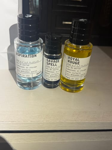 Customer's three bottles of cologne, 'Aspiration', 'Savage Spell', and 'Royal Rouge', on a wooden surface.
