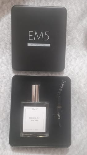 Customer's Nomade perfume bottle and a sample vial displayed inside the open, square black tin packaging.
