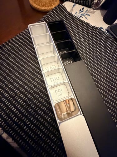 Customer's silver and black Weekly AM-PM Vitamin Cases on a table. The open silver case shows its capacity with several large pills.