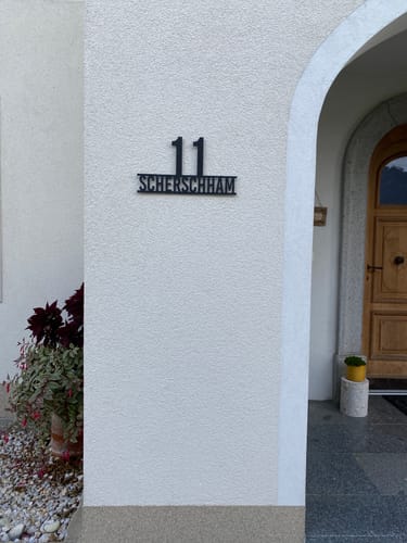 Reviewer's Stolze Nummer auf Namen in schwarz, montiert an einer hellen Hauswand neben einer Holztür.