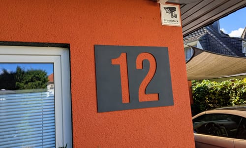 Customer's modern Hausnummernschild with number 12 mounted on an orange exterior wall next to a white window frame.