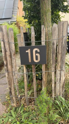Kunden-Hausnummernschild '16' in Anthrazit mit modernem Design, montiert an einem rustikalen Staketenzaun im Garten.