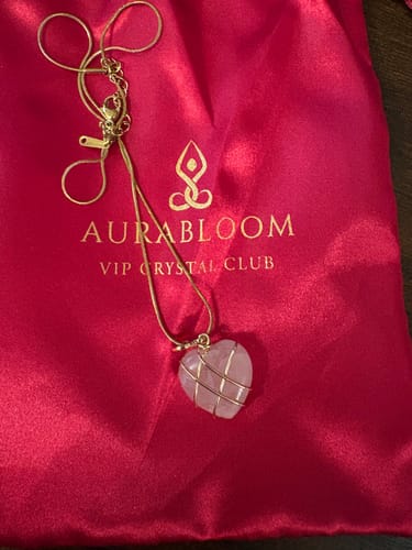 Customer's VIP Crystal Club necklace with a heart-shaped rose quartz pendant in a gold cage, on an Aurabloom pouch.