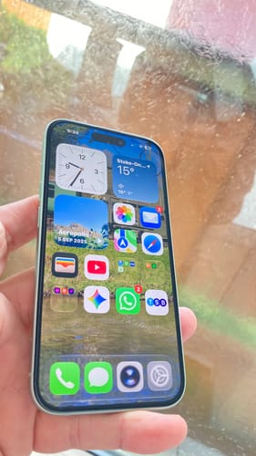 Customer's iPhone 15 in light green showing the lock screen, held by hand against a garden background.