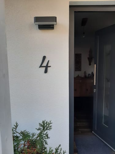 Kunden-Hausnummer Geschwungene Hausnummer montiert an einer hellen Fassade neben einer Hauseingangstür.