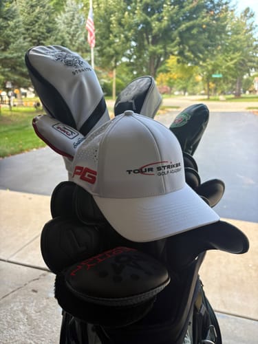 Customer's white Tour Striker hat sitting on top of golf clubs in a bag, with a blurry outdoor background.