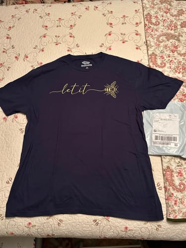 Customer's navy Let It T-Buzz T-Shirt with bee graphic lying on a floral patterned bedspread next to a shipping package