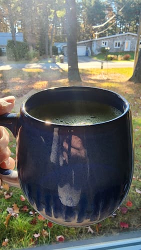 Customer's hand holding a dark blue mug of Choco Hazelnut Matcha tea in front of a sunny window.