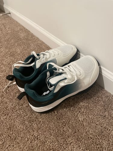 Customer's pair of white and teal CUURT Muuv court shoes with white laces, resting on a carpeted floor against a wall.