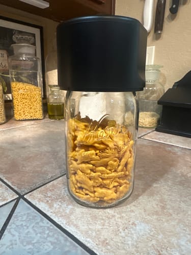 Reviewer's Mason Genie Vacuum Sealer Kit in black attached to a glass jar filled with crackers on a kitchen counter.