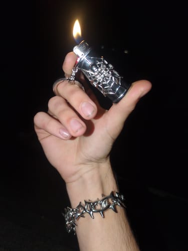 Customer's hand holding a lit silver Spider Lighter Case, wearing a detailed metal bracelet.