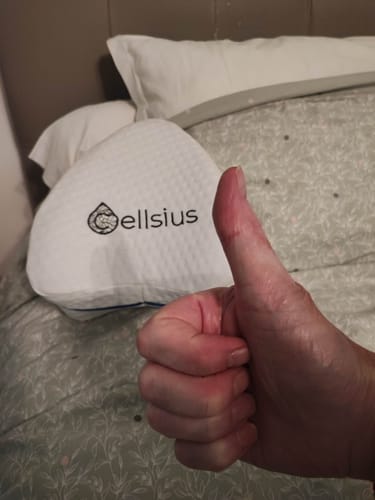 Customer's white Cellsius orthopedic knee pillow on a bed, with a hand giving a thumbs up in the foreground