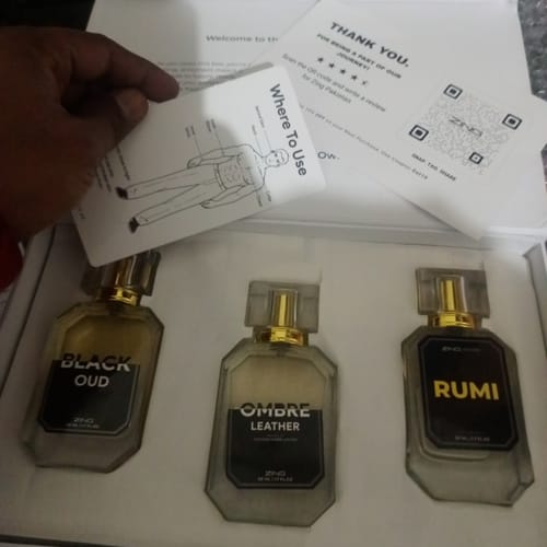 Reviewer's hand holding a card over a box with three perfume bottles: Black Oud, Ombre Leather, and Rumi.