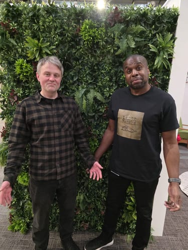 Reviewer showing a person wearing the custom black unisex t-shirt, standing with another person against a foliage wall.