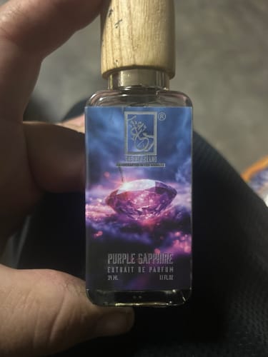A reviewer holding a bottle of Purple Sapphire perfume, showing its wooden cap and purple gem label.