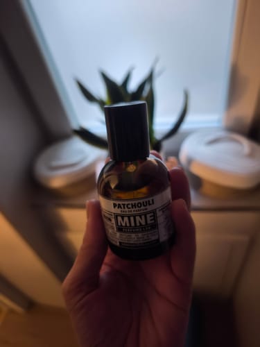 Recensore che tiene in mano la boccetta scura del profumo Patchouli in un ambiente interno.