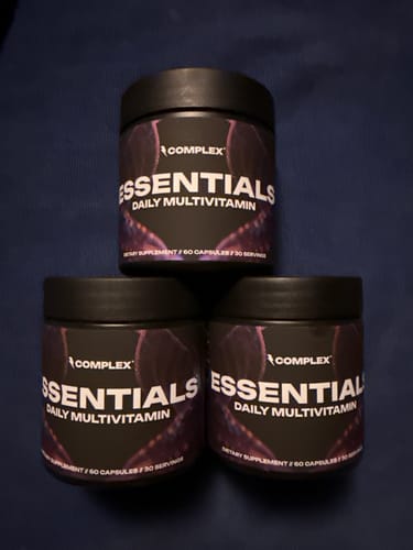 A reviewer's photo of three black tubs of Essentials Multivitamin arranged on a dark surface.