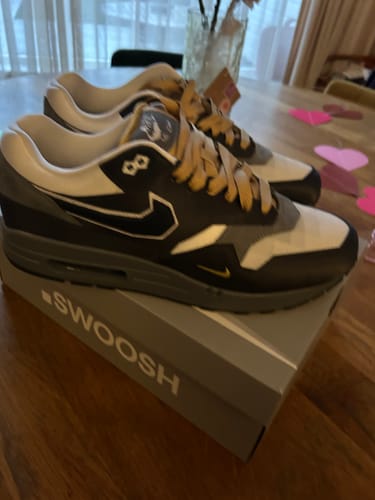 Klant toont de donkergrijze Nike Air Max 1 .SWOOSH sneakers, die op de originele schoenendoos op een tafel staan.