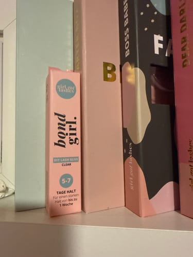 Ein Kunde zeigt die Verpackung des BOND GIRL Wimpernklebers auf einem Regal neben anderen Produkten.