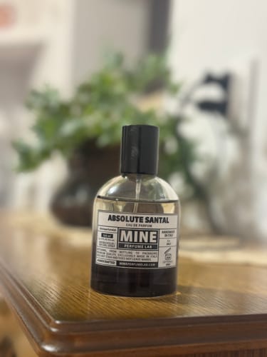 Il recensore mostra il flacone di profumo ABSOLUTE SANTAL su un mobile di legno, con una pianta sfocata sullo sfondo.