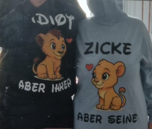 Ein Paar trägt die passenden schwarzen und grauen "Idiot Löwe" Unisex-Hoodies.