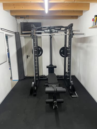 Rezensent zeigt das komplett montierte, schwarze R7-Daemon Power Rack mit Hantelbank in einem Heim-Fitnessstudio.
