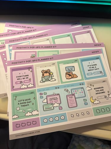 Reviewer's colorful Positivity Pop-Ups Planner Kit sticker sheets fanned out on a desk, showing cute illustrations and positive phrases.
