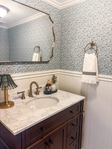 Customer's bathroom featuring the Idalie Wallpaper with a light blue floral pattern installed above white wainscoting.