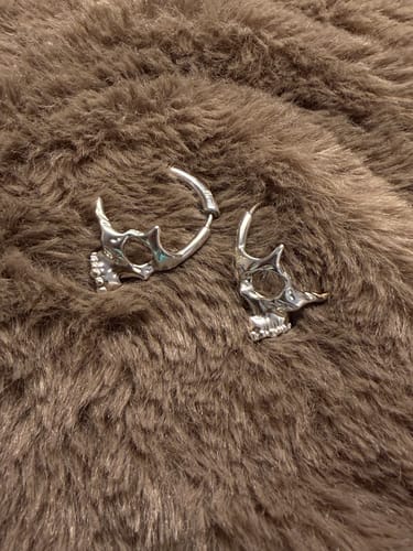 Reviewer's silver Skull Hoop Earrings on a brown fluffy surface, with one earring shown missing its back.