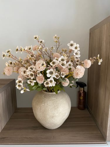 Klant toont het prachtige Wax Arrangement - Crème in een grote, getextureerde vaas op een houten plank.