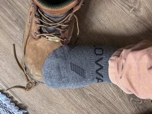 Customer's foot wearing a grey alpaca sock, shown next to a brown work boot on a wood floor.