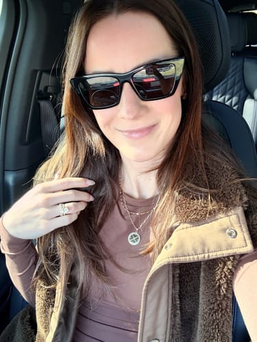Reviewer smiling in a car while wearing the Engravable Compass With Stones necklace as part of a layered look.