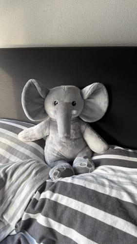 Klantfoto van de zachte Olifant Verzwaringsknuffel die rechtop op een bed zit.
