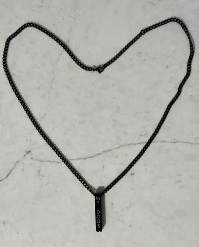 Customer's black urn pendant necklace laid in a heart shape on a marble surface. The pendant is engraved with 'MY DOGS'.