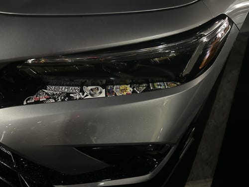 Close-up of a reviewer's 8 Year Anniversary Sticker Pack on a silver car headlight at night.