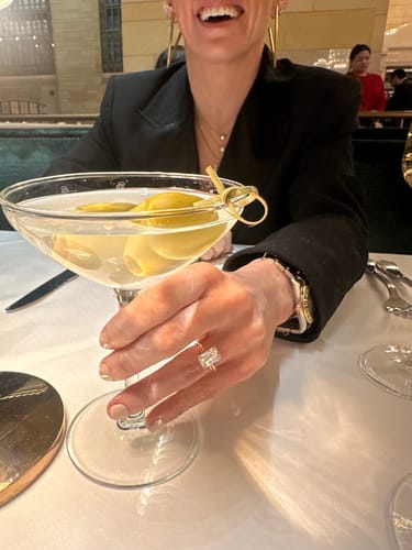 Reviewer showing off her Plunge Hidden Halo Emerald engagement ring while holding a martini glass.