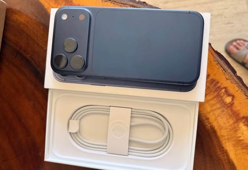 Cliente mostrando su nuevo IPHONE 17 PRO MAX azul oscuro sobre su caja abierta, con el cable visible.