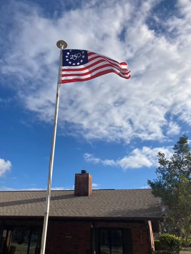 Customer's America 250™ Betsy Ross Flag waving on a flagpole against a bright, cloudy sky above a house.