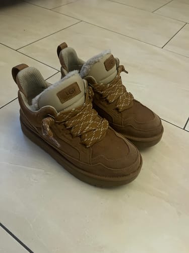 Recensent toont de kastanjebruine UGG Lowmel-schoenen met pluizige voering op een tegelvloer.