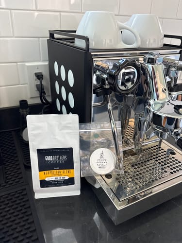 Reviewer's Espresso Paper Filters in a small container, sitting on a counter next to an espresso machine and a bag of coffee.