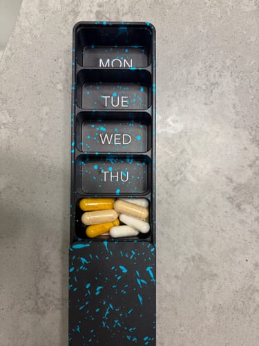 Reviewer's open black metal pill case with a blue splatter design, showing pills in a weekly compartment.