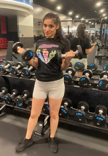 Reviewer wearing the black PowerPump Girls T-Shirt while curling two dumbbells in a gym.