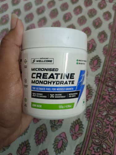 Reviewer holding the white tub of Wellcore Micronised Creatine Monohydrate in 'Kiwi Kick' flavor.