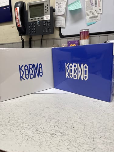 Reviewer's two K-pop albums from the lucky random box, a white and a blue version of the 'KARMA' album, on a desk at work.