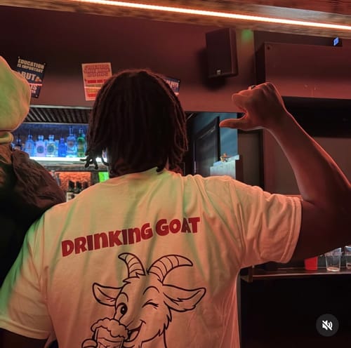 Customer from behind wearing the white unisex t-shirt, showing the 'Drinking Goat' graphic on the back in a bar.